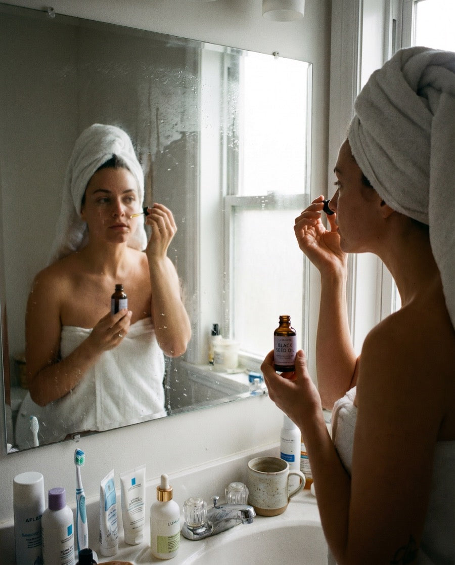 Bottle of Lumaura cold-pressed black seed oil on a bathroom shelf beside a folded towel
